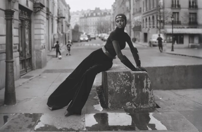 An adult female fashion model sits on concrete city street bollard in black sportswear with children playing soccer in the background.