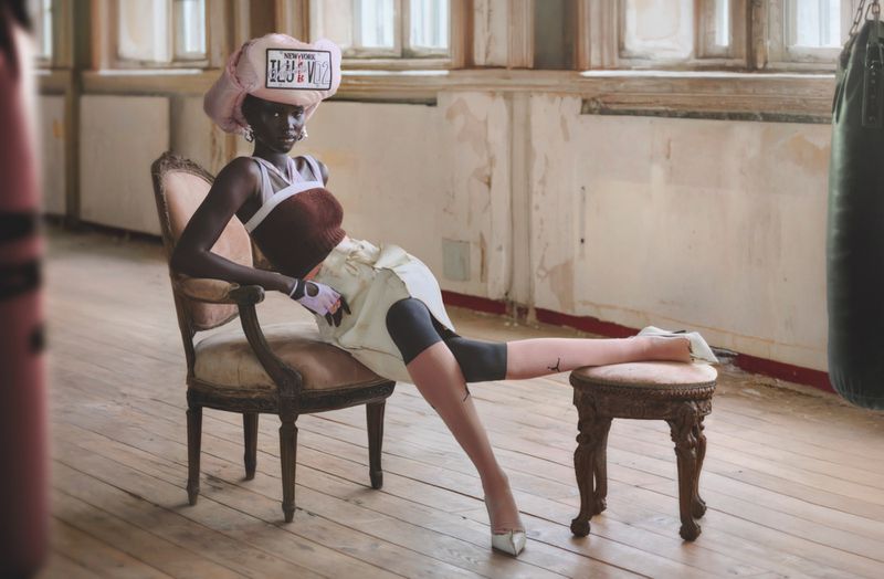 Elegant fashion photo of a woman with dark skin tone in light-colored outfit sitting in vintage chair in room with window and boxing bag.
