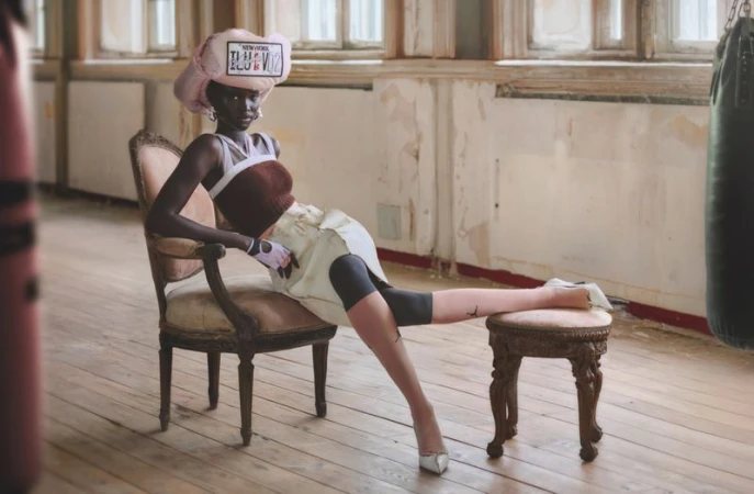 Elegant fashion photo of a woman with dark skin tone in light-colored outfit sitting in vintage chair in room with window and boxing bag.