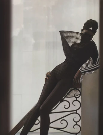 A striking fashion portrait showcases a Black female model in a black patterned bodysuit, leaning against a wrought iron railing against a bright background.
