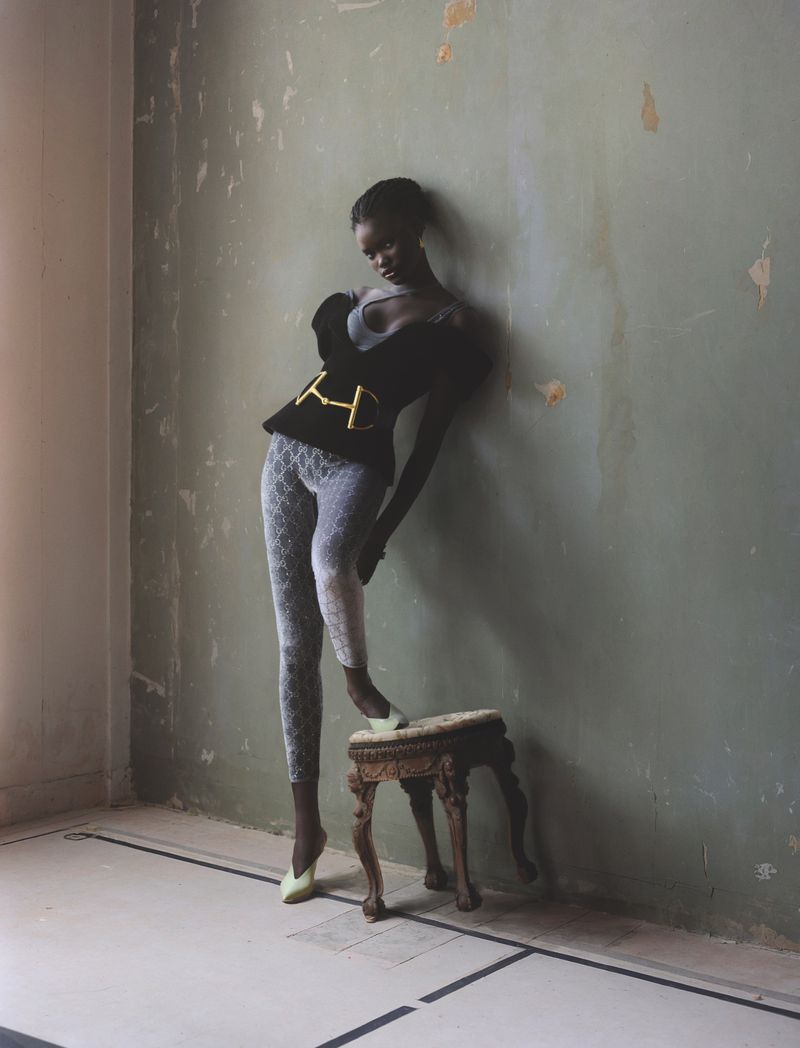 Fashion shot of a Black woman posing in a room with distressed walls. She is wearing a top with gold accents, leggings, and green shoes.