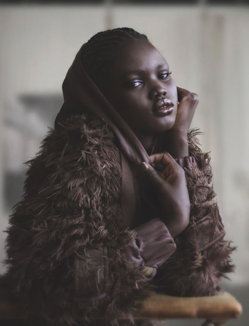 Portrait of a beautiful Black woman in a textured brown coat and hood posing indoors with neutral light and smooth skin.