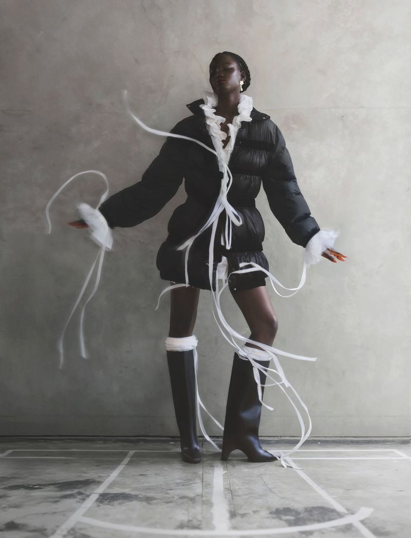 A black female fashion model wears a black puffer jacket, black boots, and white ruffle details indoors against a grey wall.