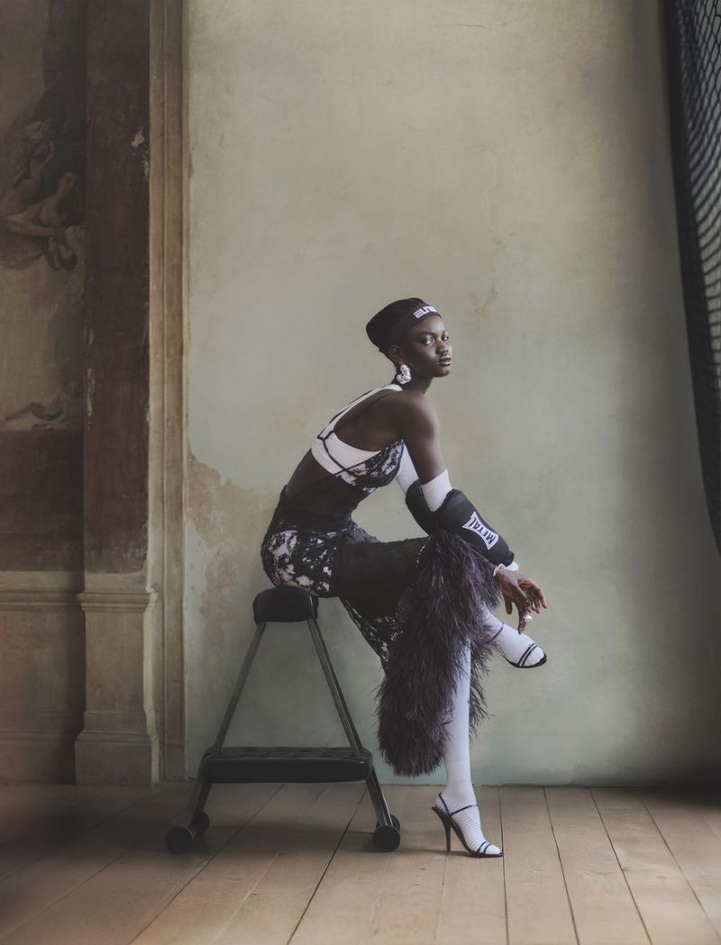 Fashion photo of a Black woman in athletic attire sitting on a step stool in an indoor setting with neutral colors and natural lighting.