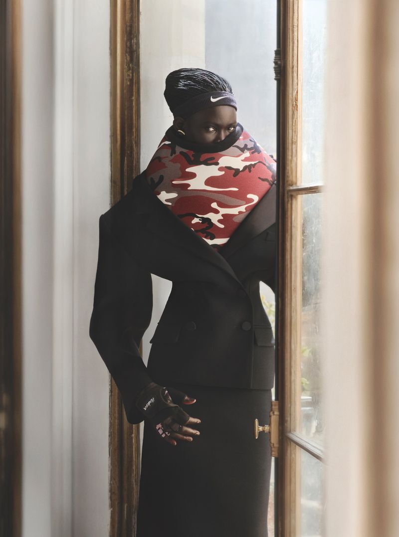 Woman in black skirt suit and red camouflage snood stands near window, looking off-camera in soft indoor light. Fashion editorial portrait.