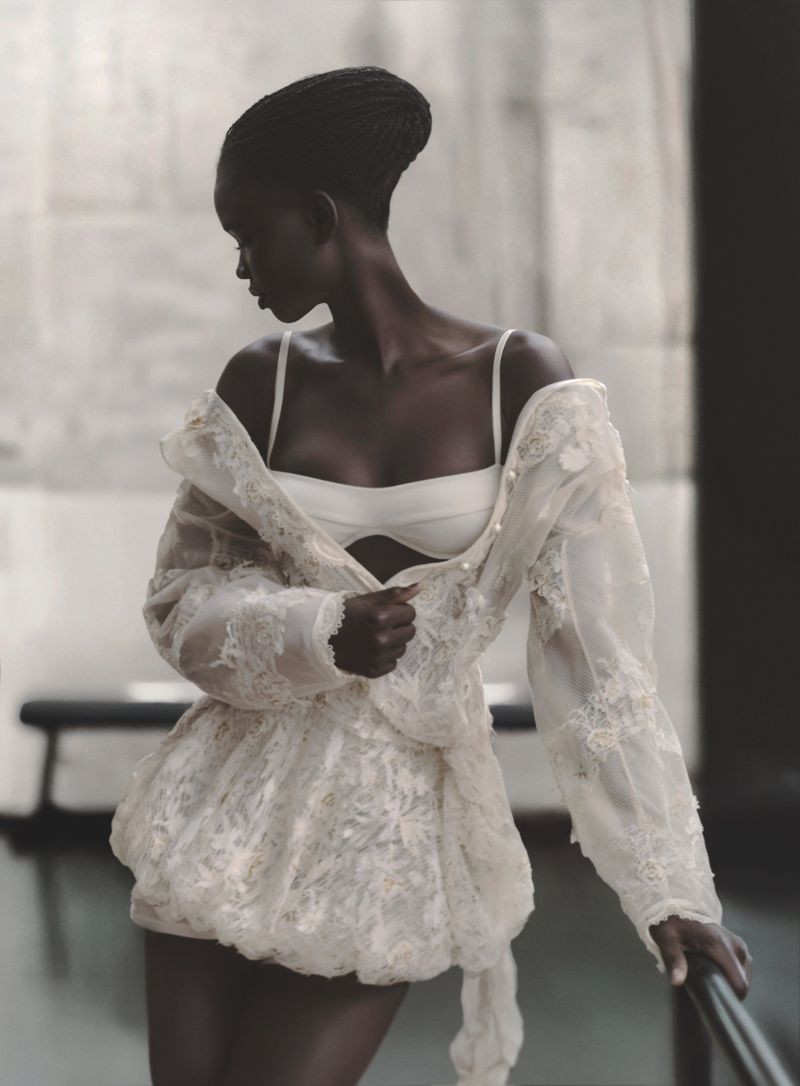 Elegant dark-skinned woman in white lingerie and a lace jacket, posing indoors with soft lighting, showcasing fashion and beauty.