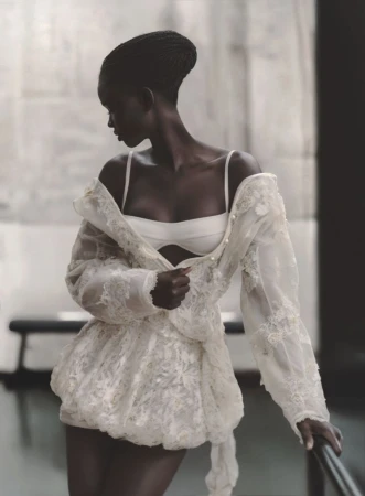 Elegant dark-skinned woman in white lingerie and a lace jacket, posing indoors with soft lighting, showcasing fashion and beauty.