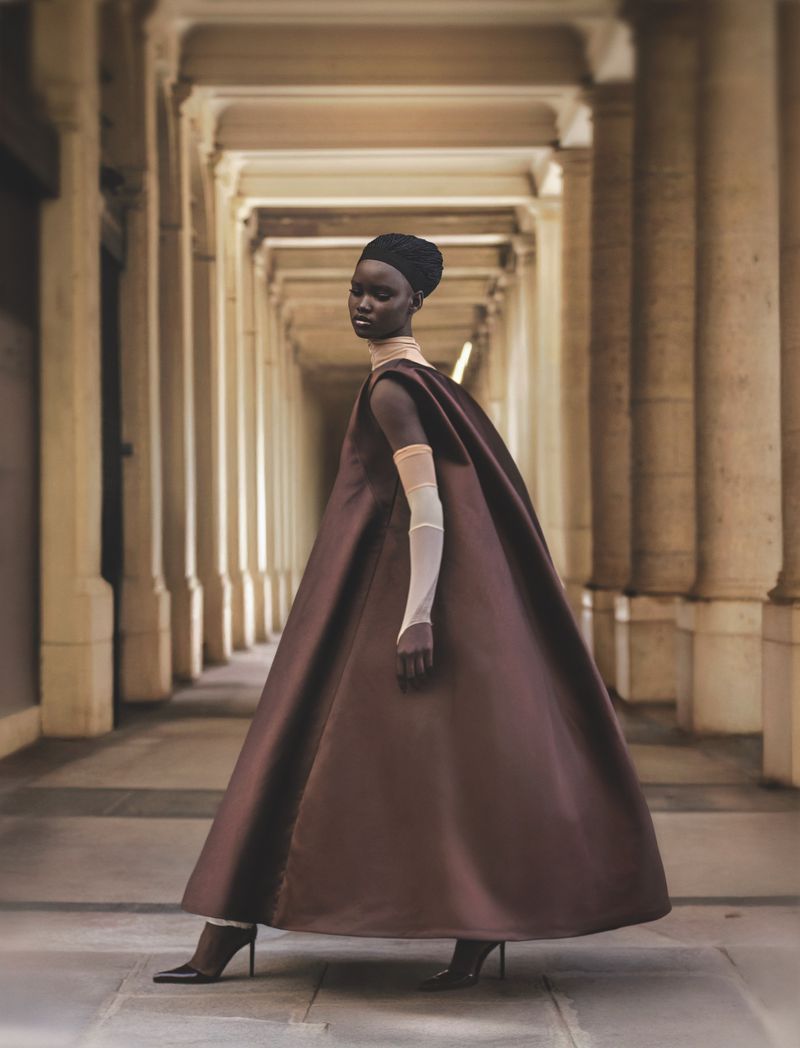 An adult female of African descent wearing a large brown dress and sheer gloves is posing in an arcade with stone pillars and arches.
