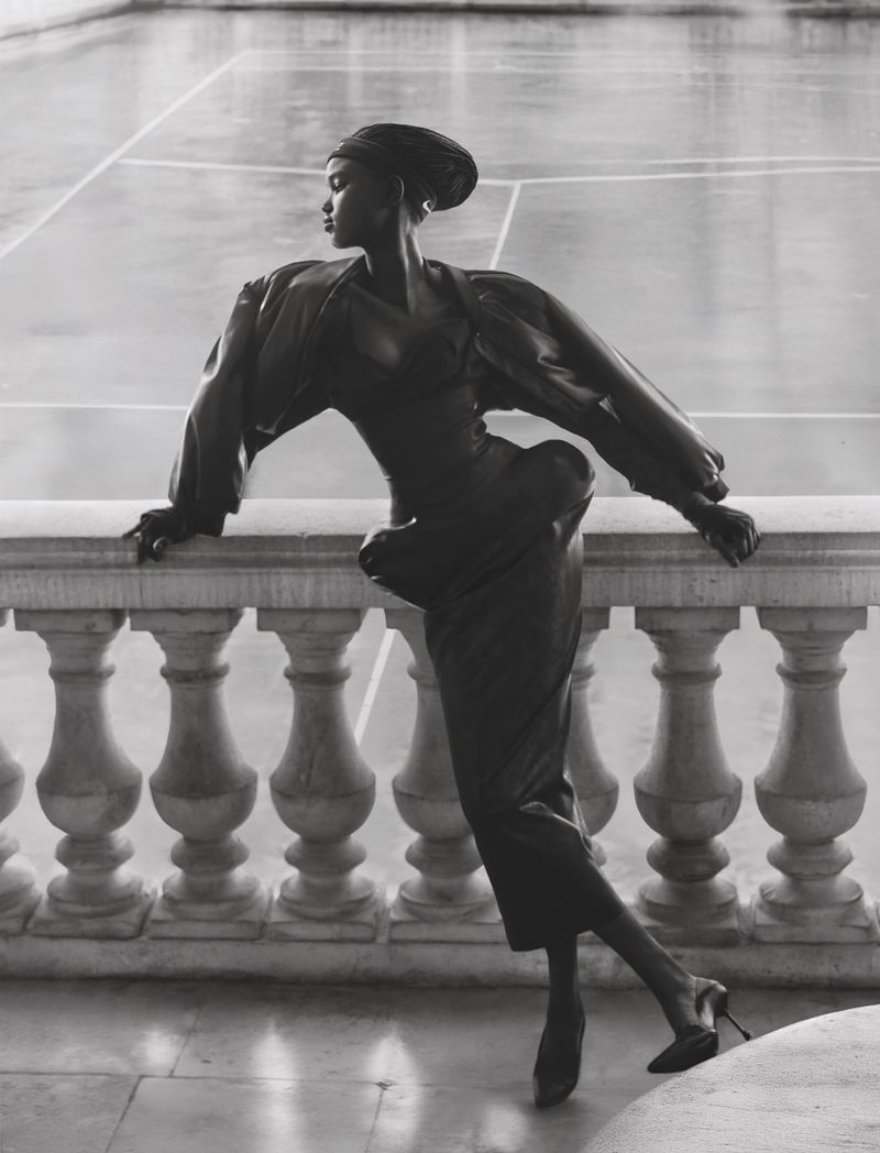 Elegant woman in dark couture dress leaning on a white stone balustrade, set against a neutral architectural backdrop.