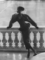 Model Nyakong Chan in a sculptural black dress leaning against a stone balustrade above a tennis court in black and white.