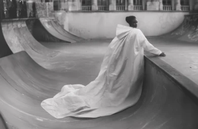 Model Nyakong Chan wears an expansive hooded white cape gown flowing down a concrete skate park ramp in black and white.