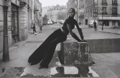 Model Nyakong Chan wearing an all-black ensemble and headwear poses leaning against a concrete block on an urban street.
