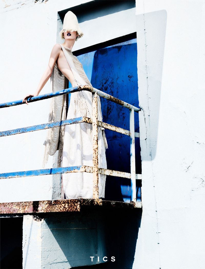Model in a white dress and hat stands on a rusted balcony with a bright blue door in the background in a fashion-style outdoor shot.