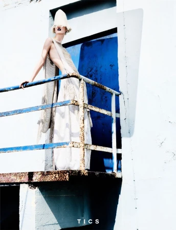 Model in a white dress and hat stands on a rusted balcony with a bright blue door in the background in a fashion-style outdoor shot.
