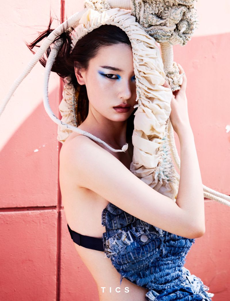 A young adult woman poses against a pink wall wearing a blue denim garment and a rope-like headdress for a fashion shot.
