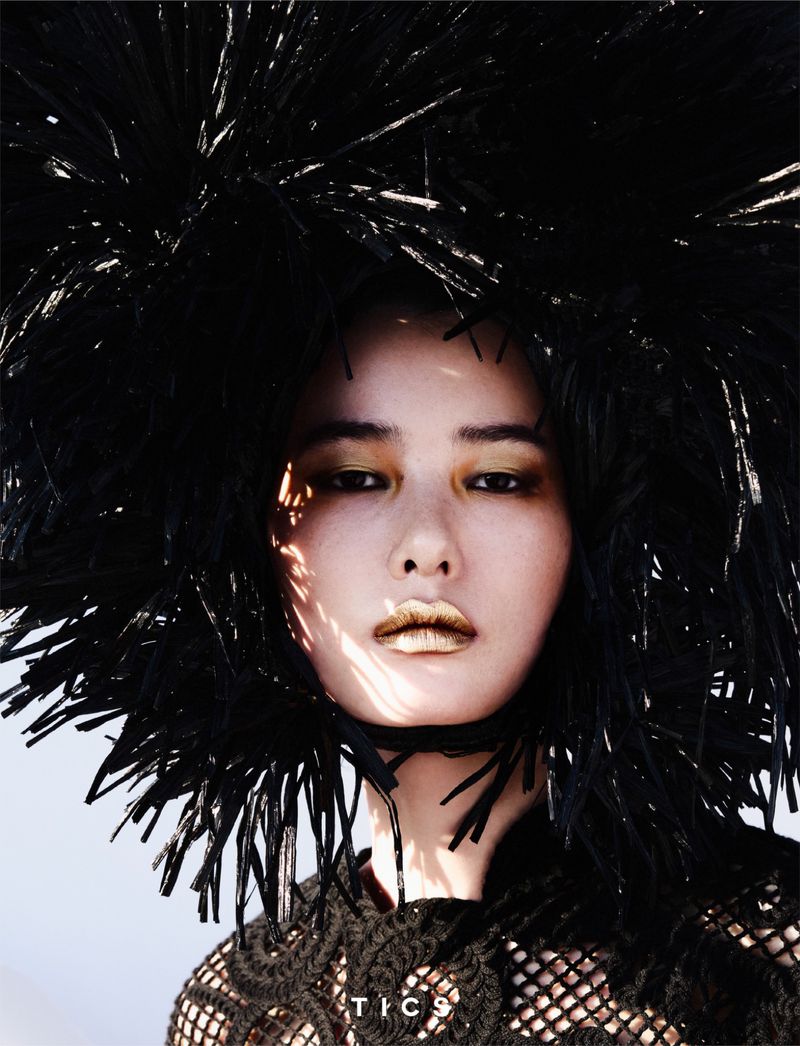 Striking portrait of a woman with gold makeup and a black feathery headpiece, set against a stark white background, showcasing high-fashion editorial style.