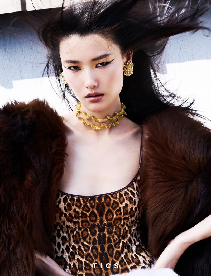 Asian woman poses in a leopard-print dress with gold jewelry and a brown fur coat, striking a fashionable pose outdoors.