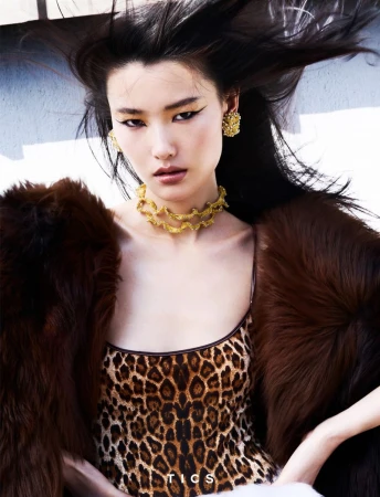 Asian woman poses in a leopard-print dress with gold jewelry and a brown fur coat, striking a fashionable pose outdoors.
