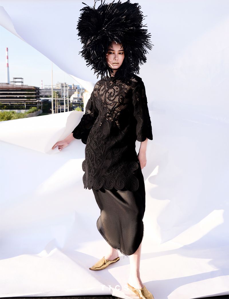 An Asian woman is posing in a black lace dress and a large feather hat in a studio with a white backdrop, and city buildings visible.