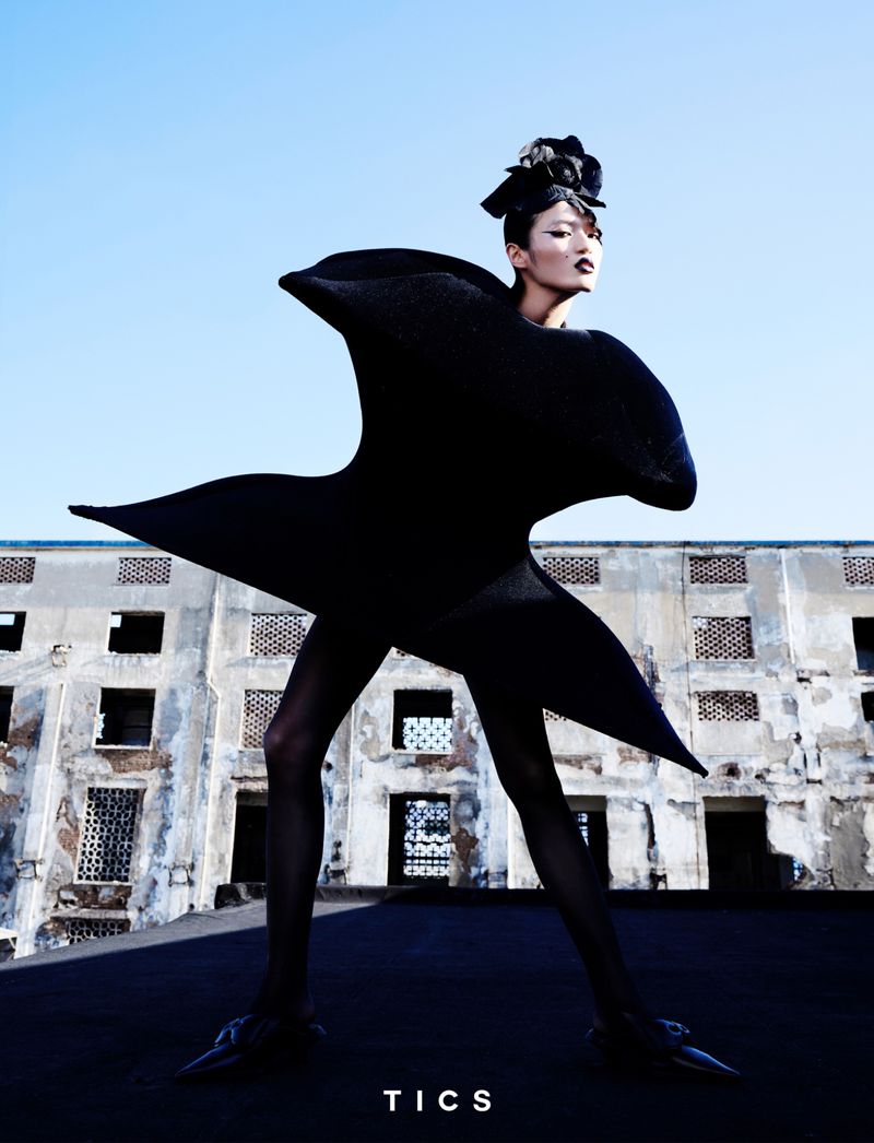 Woman in avant-garde black dress with headpiece poses on rooftop against a weathered building and blue sky in a fashion editorial style.