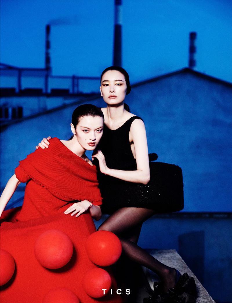 Two Asian women in a high-fashion photoshoot, one in black, one in red, posing on concrete against a blue industrial background with chimneys.