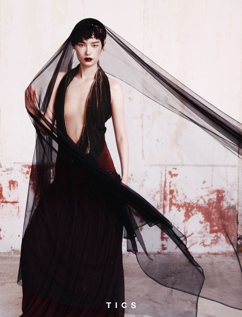 Fashion photo features a solo adult woman with dark hair in a long black dress and sheer black veil, posing against a distressed wall.