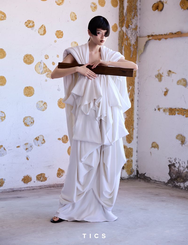 Full shot of a woman in a white ruffled dress holding a wooden plank indoors against a distressed wall with circular paint patterns.