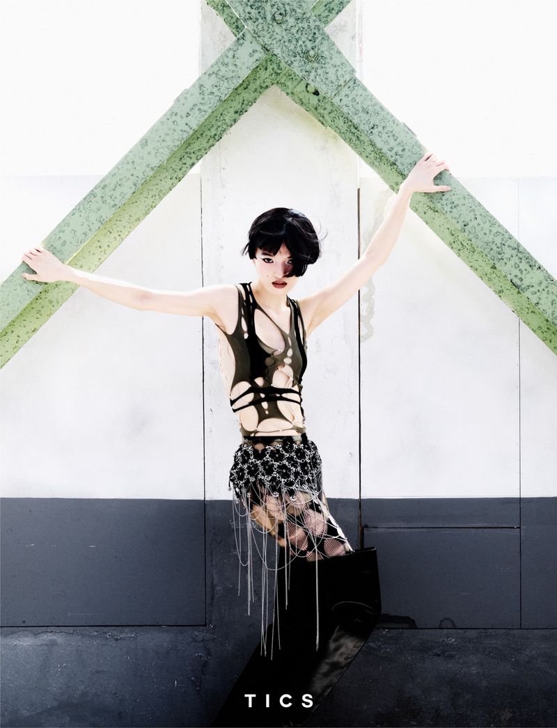 Young woman in a sleeveless black cutout top, decorated skirt, and knee-high boots poses on a platform in front of a concrete wall.