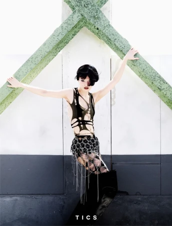 Young woman in a sleeveless black cutout top, decorated skirt, and knee-high boots poses on a platform in front of a concrete wall.