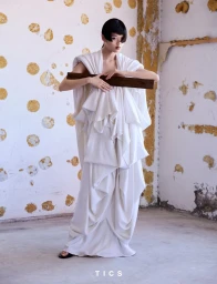 Model with short black hair wearing a voluminous draped white gown and holding a dark wooden plank in a distressed room.