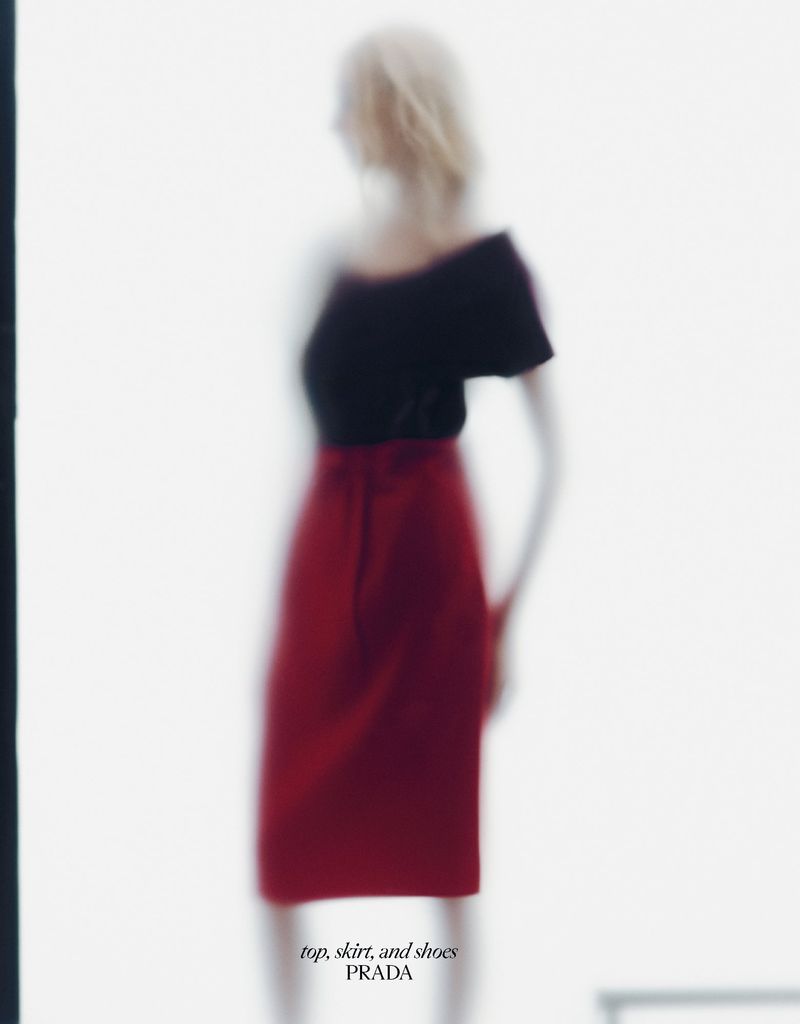 Fashion shot of a blonde model wearing a black top and red skirt against a blurred white backdrop with the Prada logo.