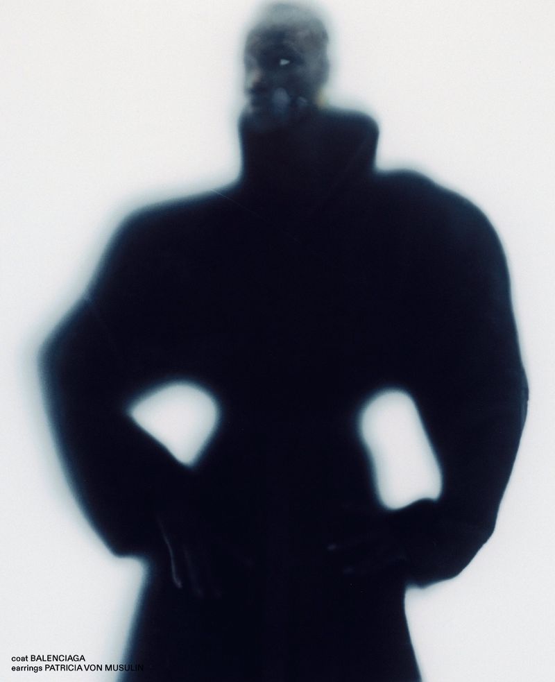 Blurred silhouette of a model wearing an exaggerated black coat with hands on hips against a luminous white background.
