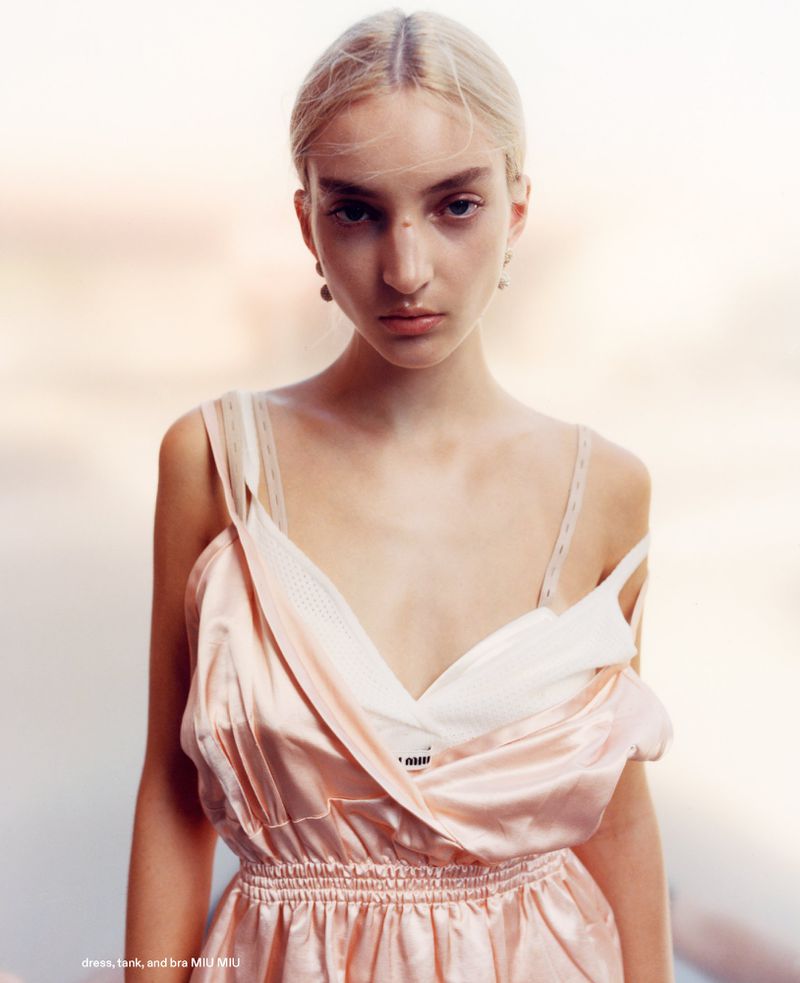 A young woman with fair skin and blonde hair poses in a pink satin dress with a white underlay, against a blurred light background.