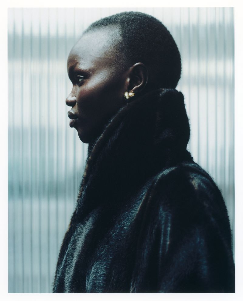 Profile portrait of a woman with dark skin and a shaved head, wearing a fur coat against a grey, corrugated metal wall indoors.