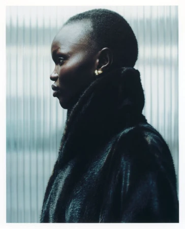 Profile portrait of a woman with dark skin and a shaved head, wearing a fur coat against a grey, corrugated metal wall indoors.