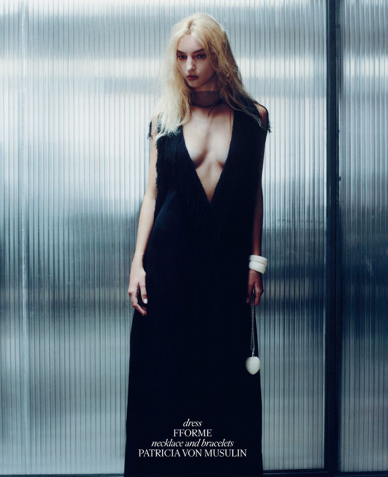 Blonde woman in black dress poses against a corrugated metal wall, showcasing a necklace and bracelets in a fashion editorial shot.