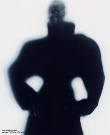 Blurred silhouette of a model wearing an exaggerated black coat with hands on hips against a luminous white background.