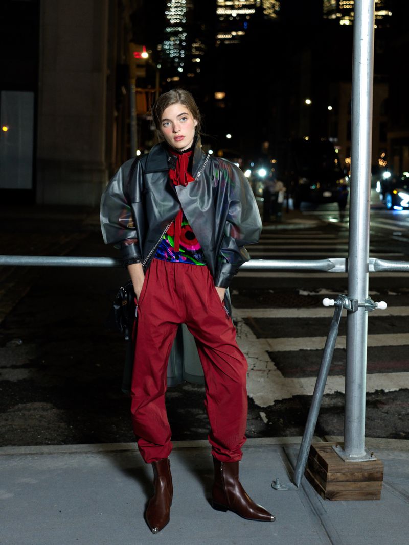 A young woman in a black coat and red pants stands on a city street at night with buildings and traffic in the background.
