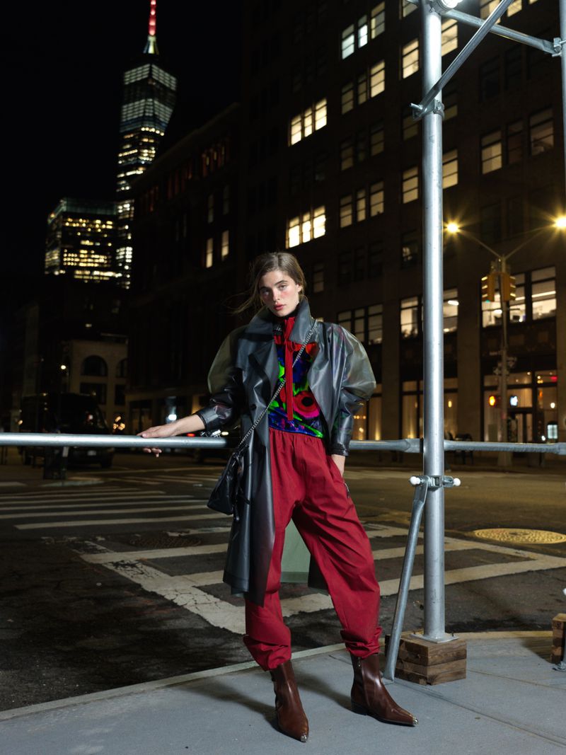 Fashion model in a grey transparent trench coat and red pants poses at night in an urban setting with buildings in the background.