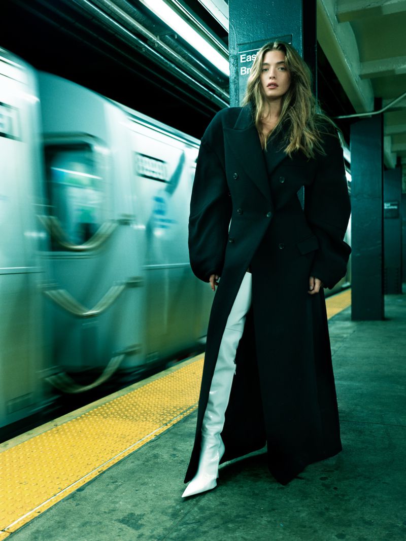 Fashion shot of a woman in a black coat and white boots standing on a subway platform, with a blurred train in the background.