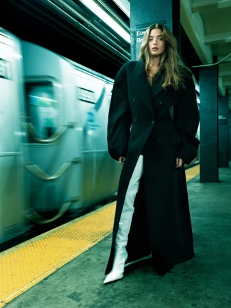 Fashion shot of a woman in a black coat and white boots standing on a subway platform, with a blurred train in the background.