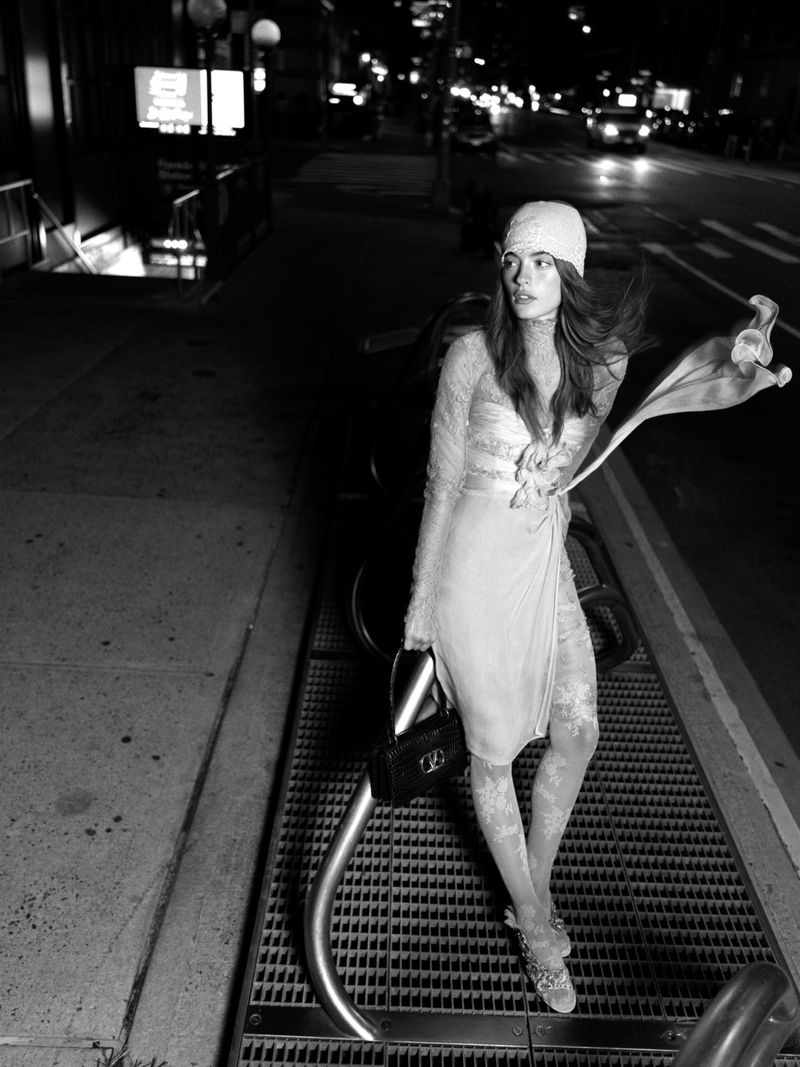 Monochrome portrait of a woman in a dress and beanie standing on a city sidewalk at night, holding a purse with cars in the distance.