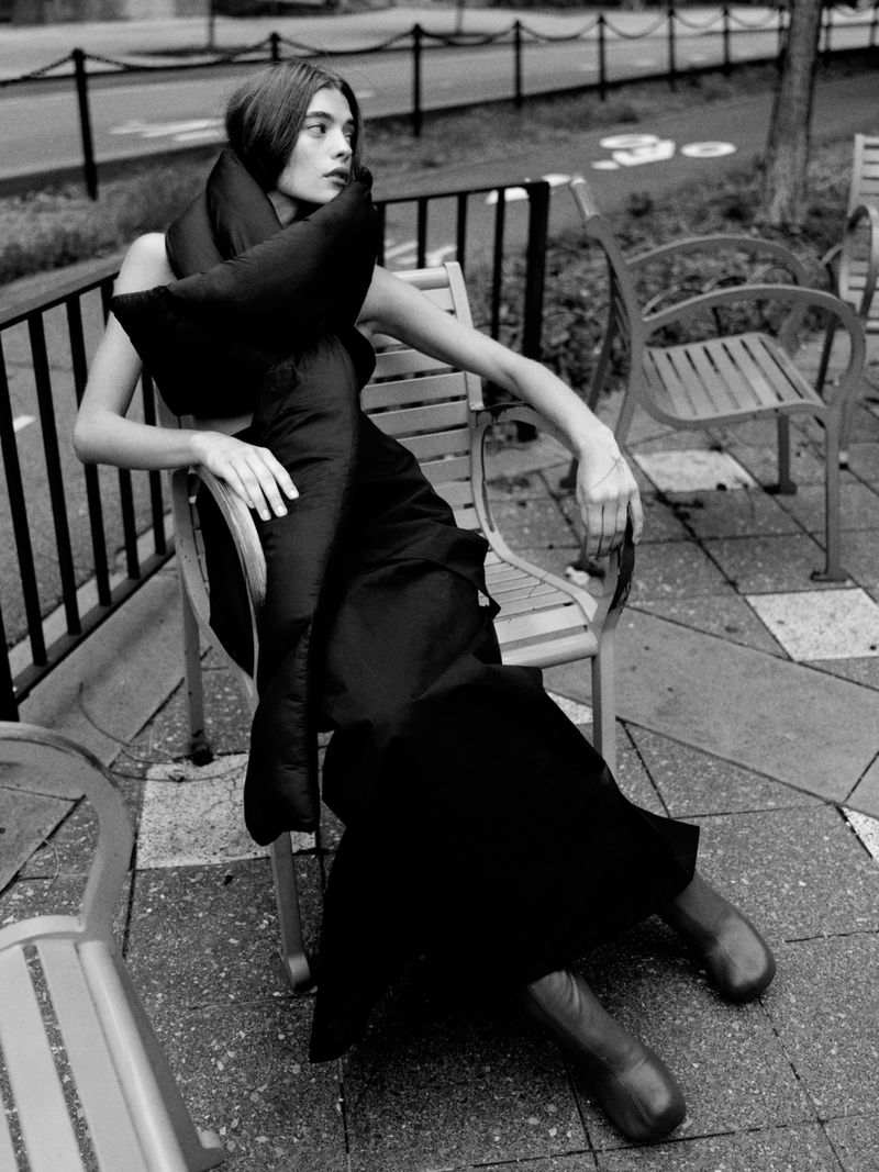 A woman poses in a black dress and scarf on a metal chair outdoors with trees in the background in a monochrome fashion shot.