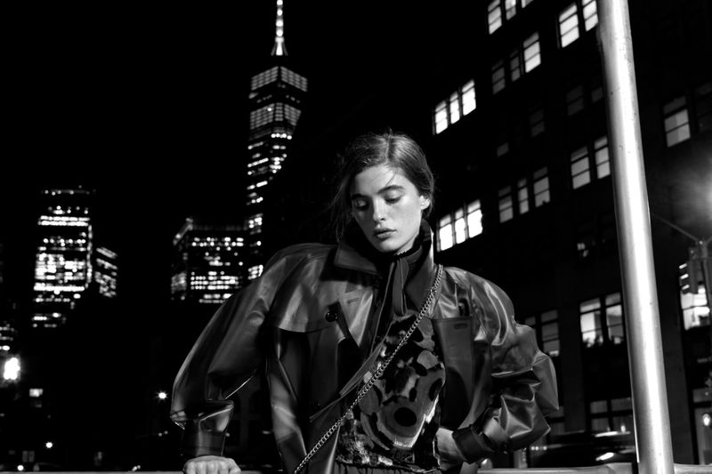 Black and white portrait of a woman wearing a jacket in front of city buildings at night with illuminated windows and street lights.