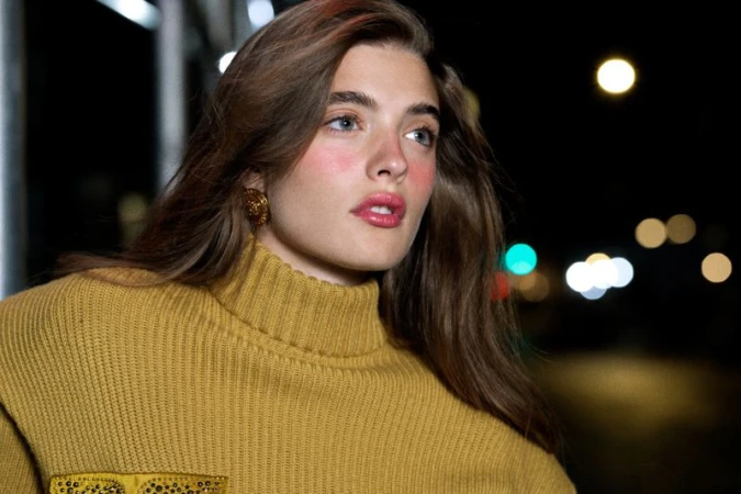 Portrait of woman with long brown hair wearing gold-colored turtleneck sweater, posing at night with bokeh lights in background.