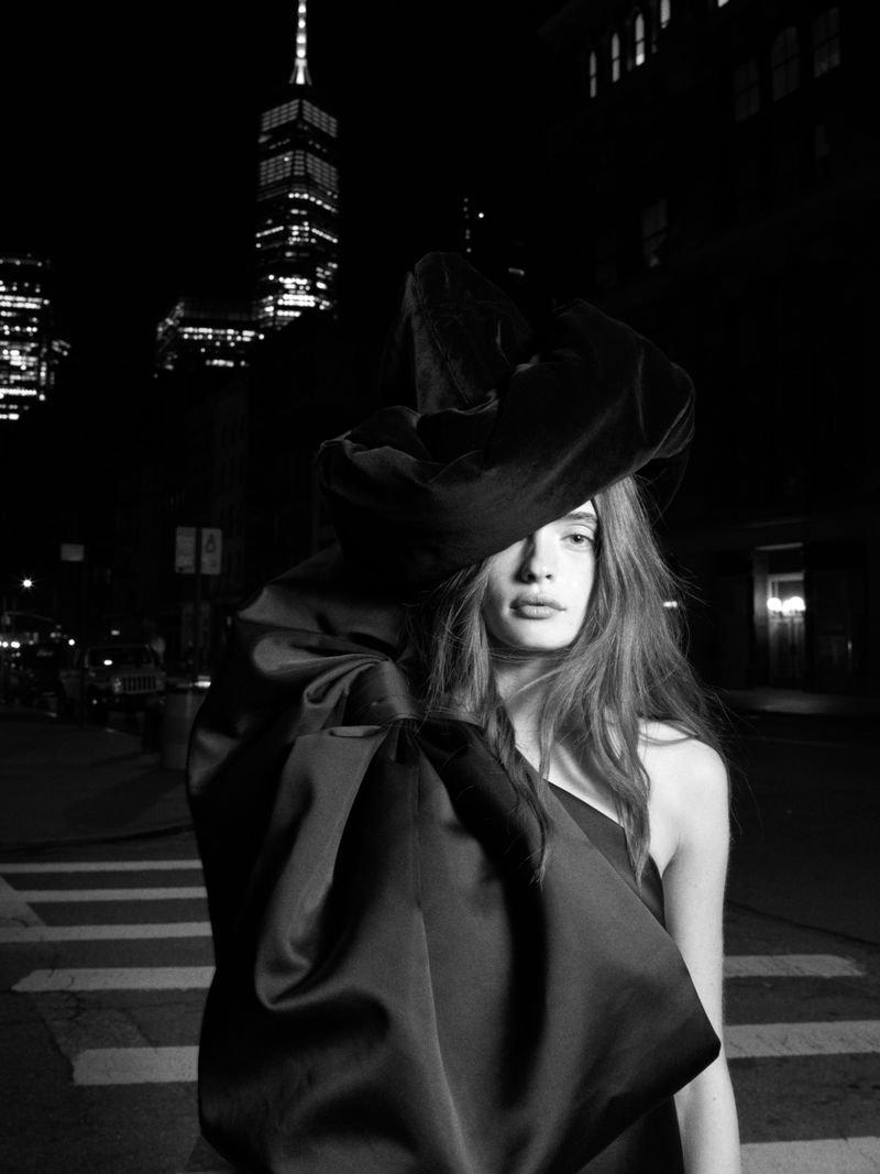 A model in a large black dress stands on a city street at night with a tall building in the background. Fashion editorial shot.