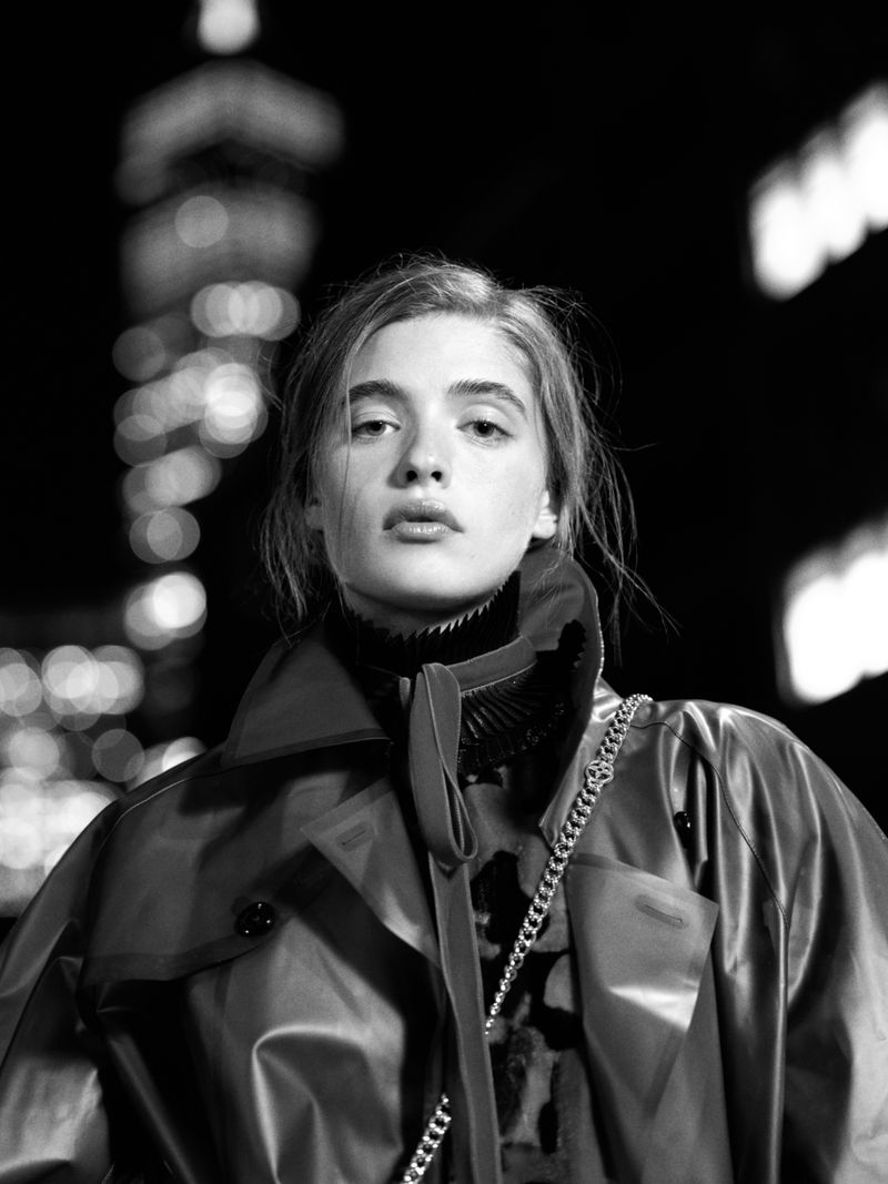 Black and white portrait of a woman in an oversized coat and pleated collar against city night lights.