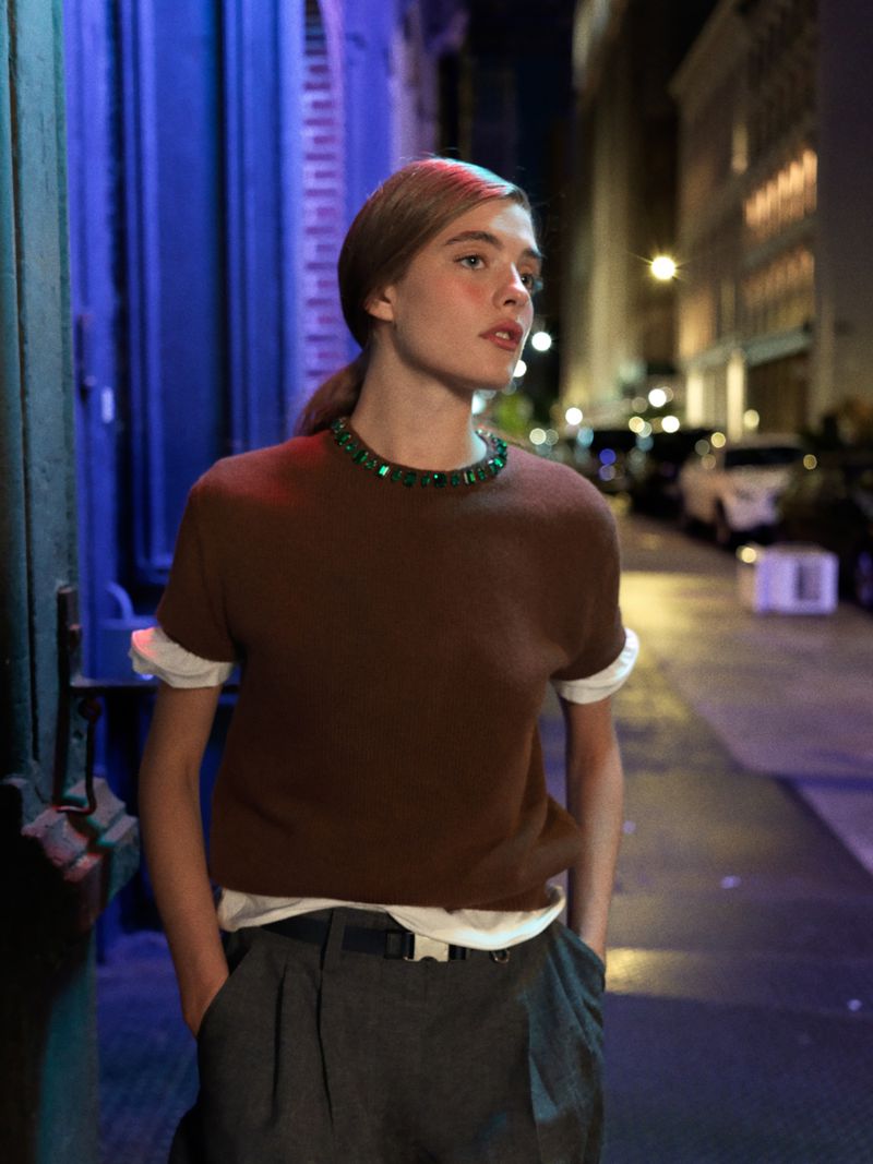 A young adult female poses in an urban environment at night, wearing a brown shirt, grey slacks, and a green beaded necklace.