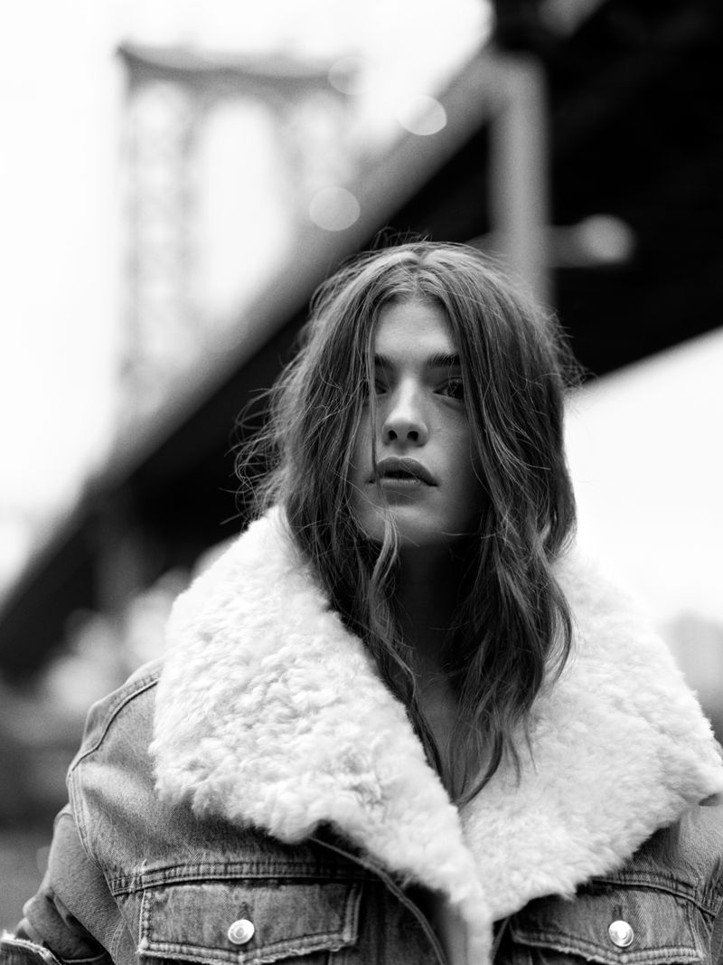 Black and white portrait of a woman with long hair wearing a denim jacket in front of a bridge.