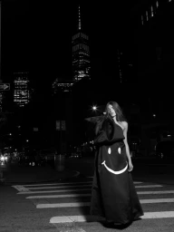 Model wearing a voluminous black gown with a bold white smiley face graphic stands in an urban crosswalk at night.